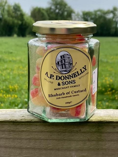 A glass jar of A.P. Donnelly and Sons Rhubarb and Custard sweets placed on a wooden surface with a natural background.