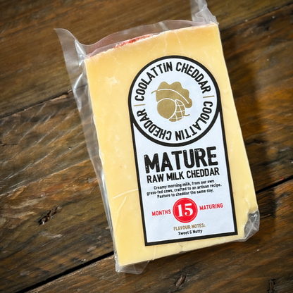 A packaged piece of Coolattin Mature Raw Milk Cheddar cheese on a wooden surface with a label indicating it is matured for 15 months