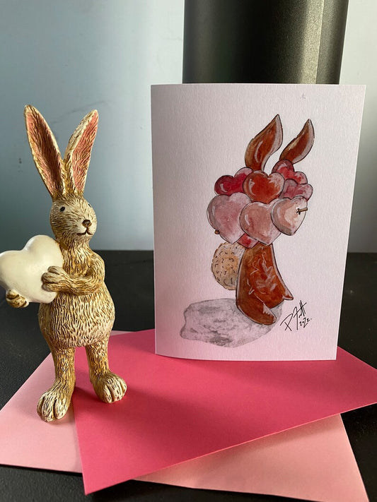 A greeting card depicting a rabbit holding a berry, with a background of a dining table and a piece of paper with a drawing of a rabbit holding a heart.