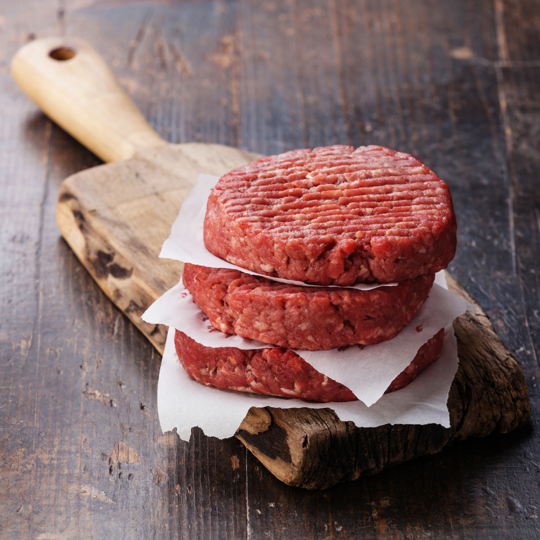 Three raw ground beef burger patties on top of a wooden cutting board, wrapped in plastic